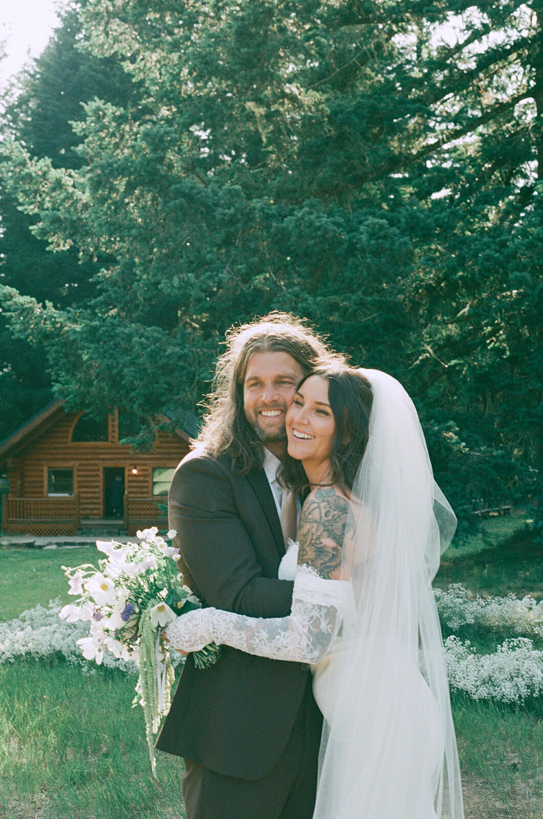 Smiling bride and groom portrait in front of rustic cabin with bouquet by Kvetka Flower Portland OR