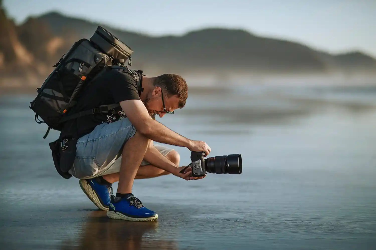 Man with a backpack squatting by a body of water, holding a camera.