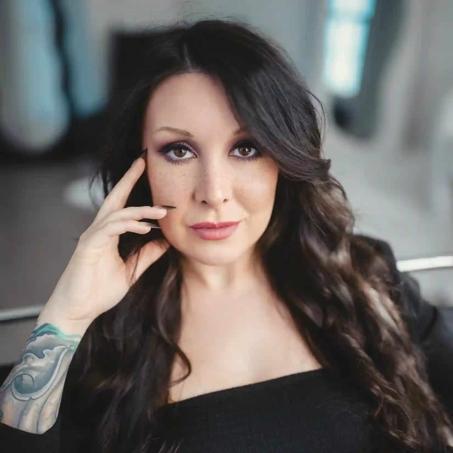 Woman with tattoos on her arm touching her face in a blurred indoor setting