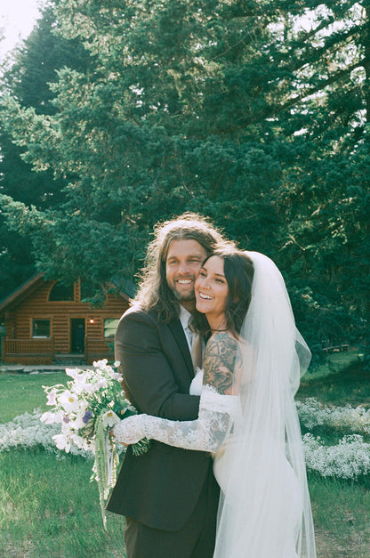 Smiling bride and groom portrait in front of rustic cabin with bouquet by Kvetka Flower Portland OR