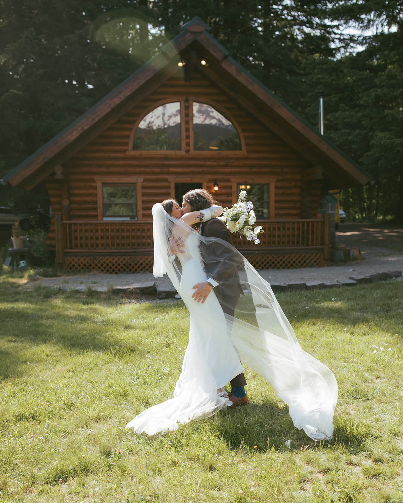 Bride and groom kissing in front of rustic cabin with bouquet designed by Kvetka Flower Portland OR