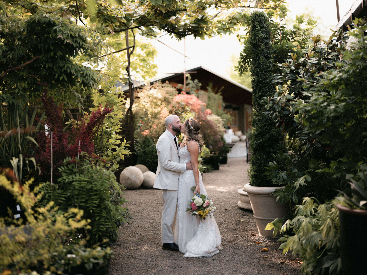 Wedding couple standing in a garden setting with lush greenery and a rustic building in the background.