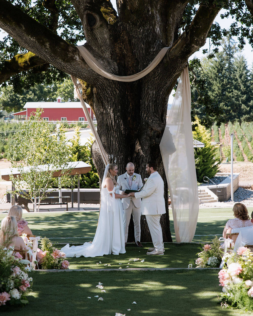 Bride and groom during their outdoor ceremony under a large oak tree decorated with flowers by Kvetka Flower.