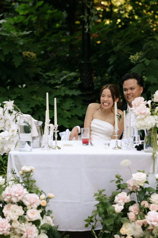 Bride and groom laughing at sweetheart table surrounded by lush floor arrangements of roses and orchids by Kvetka Flower at McMenamins Edgefield