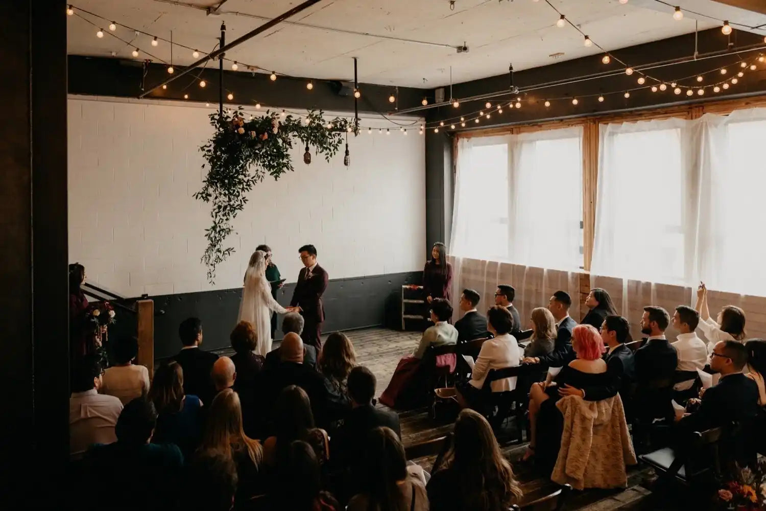 Wedding ceremony taking place in a modern indoor setting with string lights and large windows.